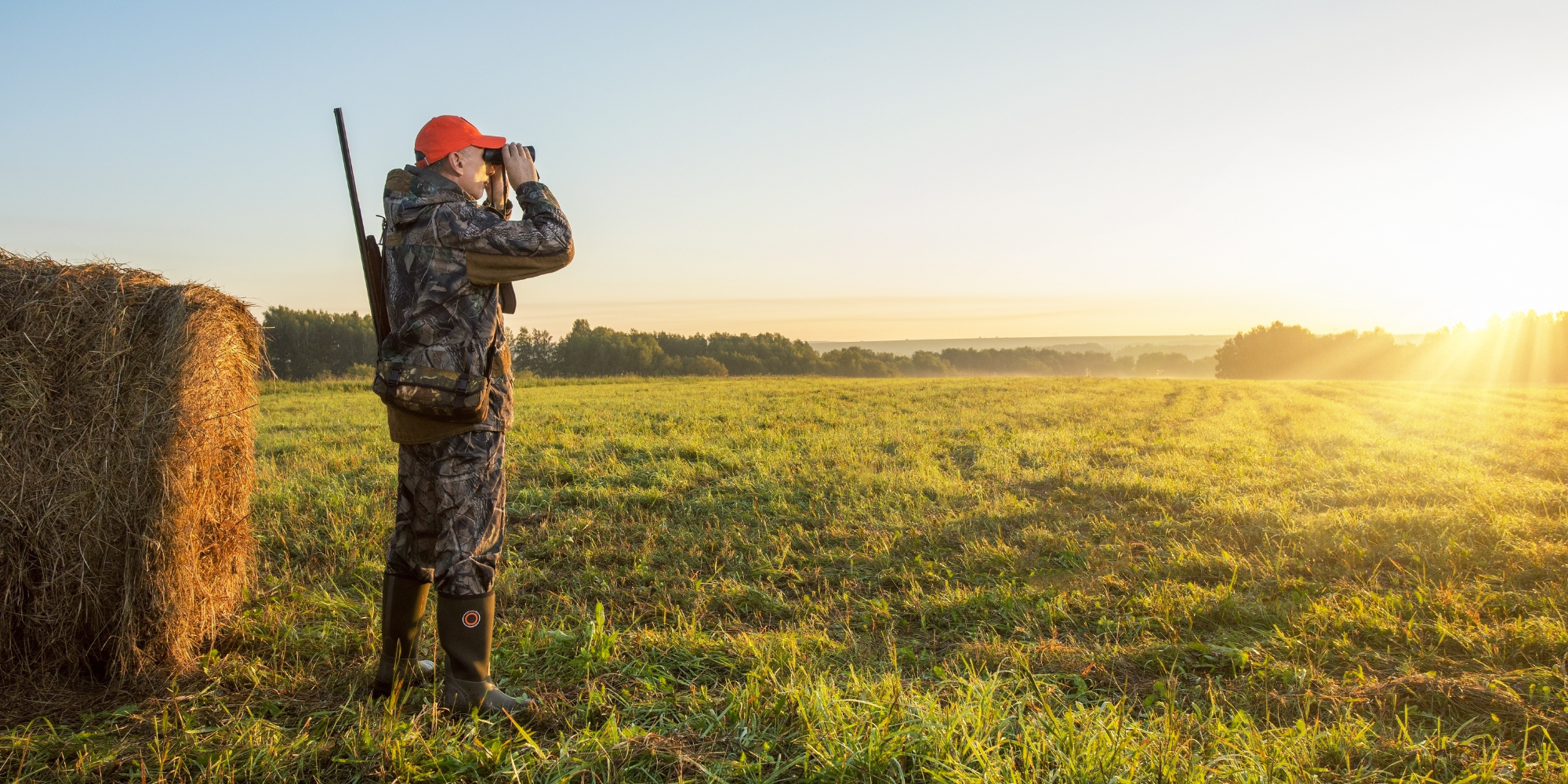 A hunter is positioned in a field, adjacent to a hay bale, embodying the spirit of outdoor exploration and wildlife pursuit.