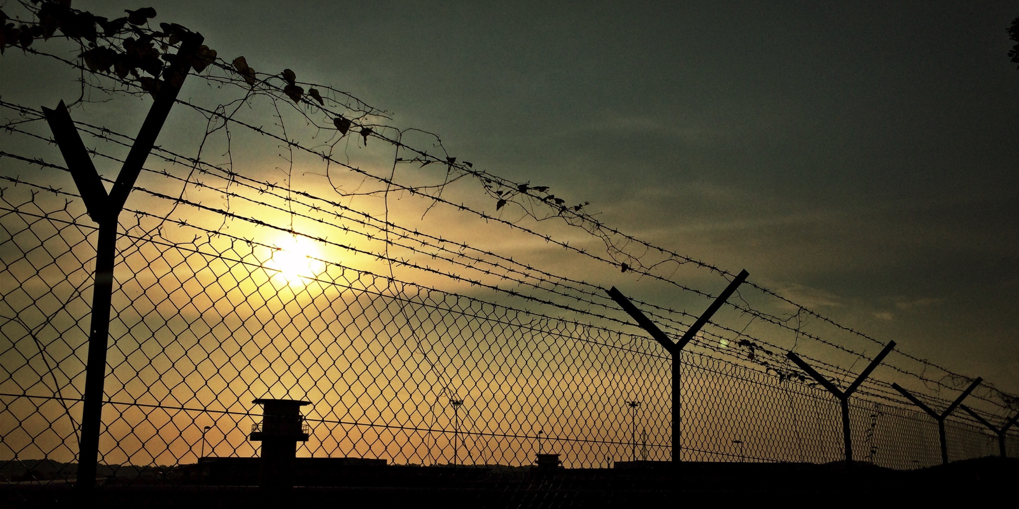 A chain-link fence topped with barbed wire is silhouetted against a sunset sky.